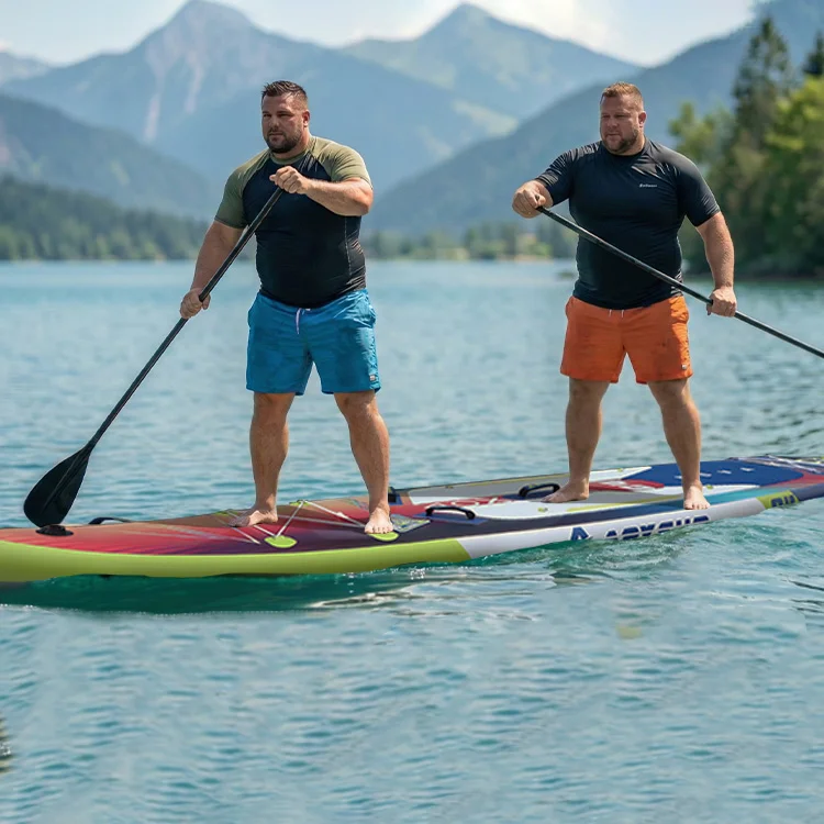 Two men paddleboarding on a colorful board on a lake with mountain backdrop, wearing colorful shorts and holding paddles.
