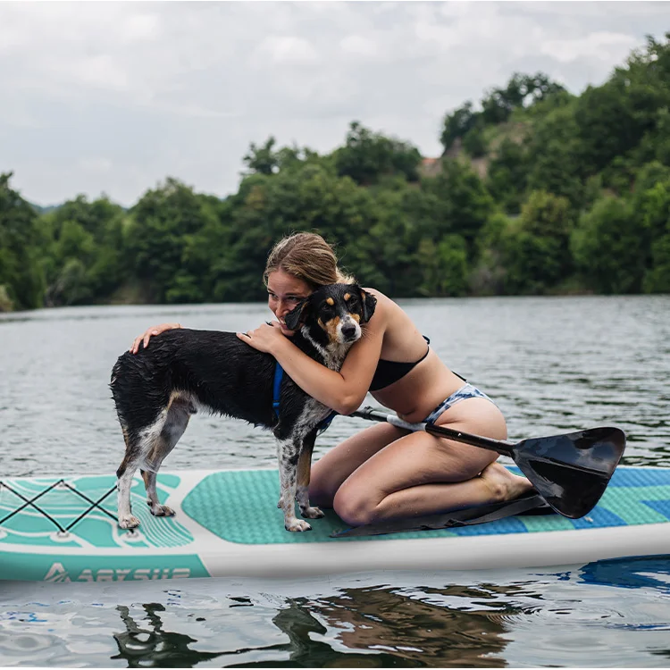 Woman in swimwear hugging a dog on a paddleboard in a calm lake with lush green trees in the background.