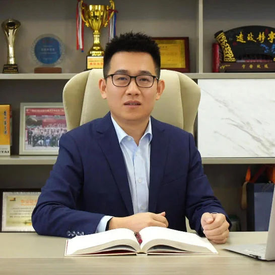 Businessman seated at a desk with an open book in a professional office setting, surrounded by shelves displaying trophies and awards.