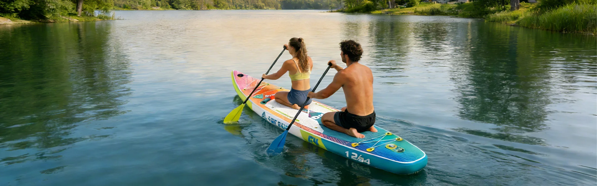 A couple paddleboarding on a calm lake surrounded by trees. They are seated on a colorful board under clear skies.