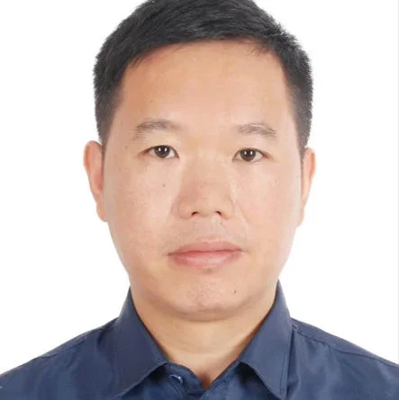 Individual in a blue collared shirt with short black hair against a white background.