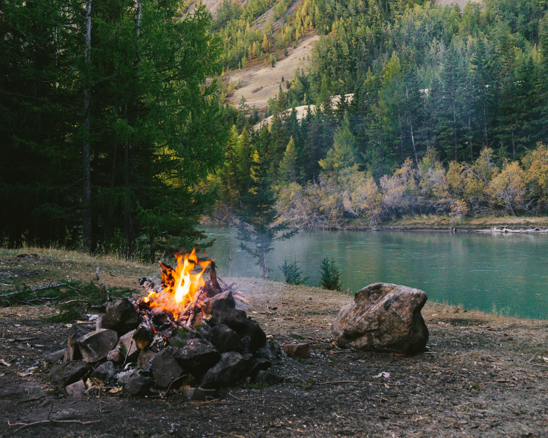 A campfire burning on rocky ground near a calm river surrounded by dense forest and hills.