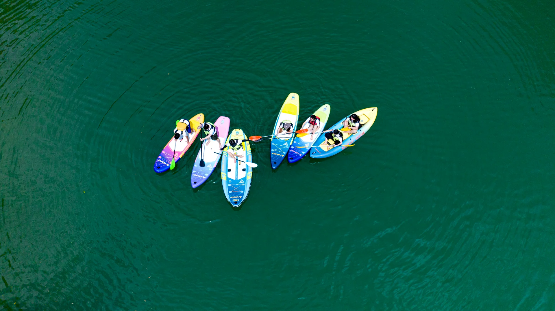 Aerial view of a group of people on colorful paddleboards on a calm lake. The boards are arranged in a circle, and paddlers are wearing life jackets.