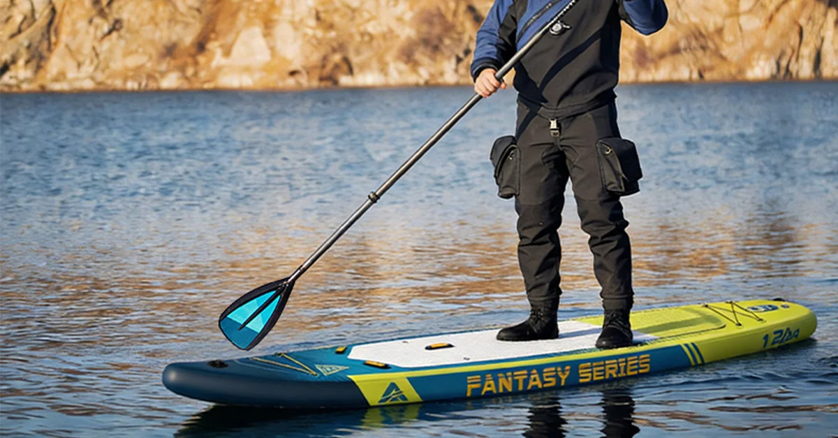 Person standing on a paddleboard labeled 'Fantasy Series' in a lake with rocky cliffs, using a paddle.