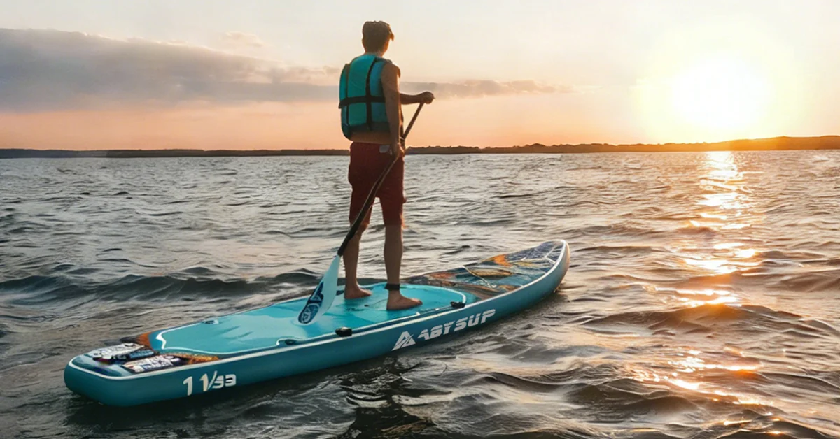 Person paddleboarding on a lake at sunset, wearing a teal life vest and standing on a blue paddleboard with gentle waves.