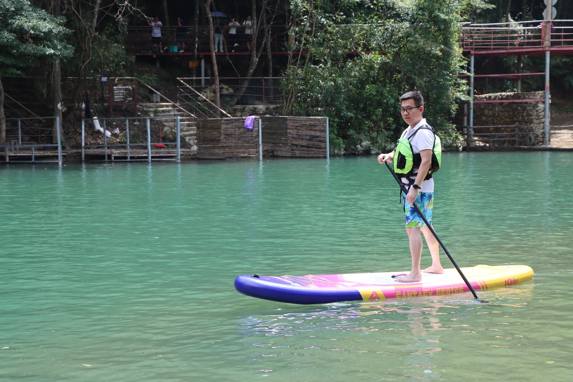 Individual standing on a colorful paddleboard in a lake, wearing a safety vest and holding a paddle. Lush vegetation in the background.