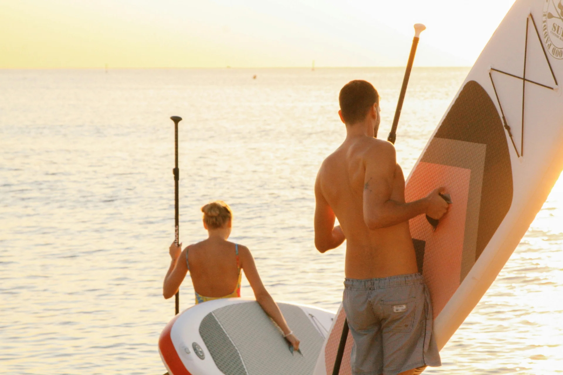 A couple preparing to paddleboard on the ocean at sunset, holding paddles and boards with the sun setting over the horizon.