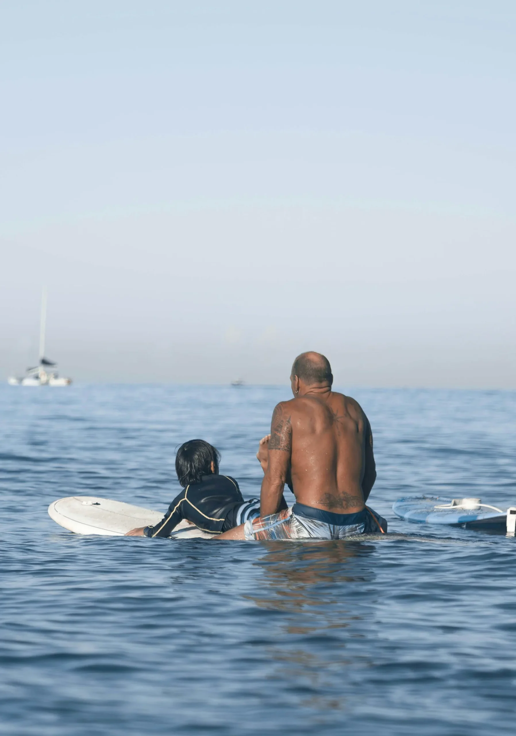 A man and child sitting on surfboards in the ocean, with a distant sailboat visible on the horizon. The man has tattoos and the child is wearing a wetsuit.