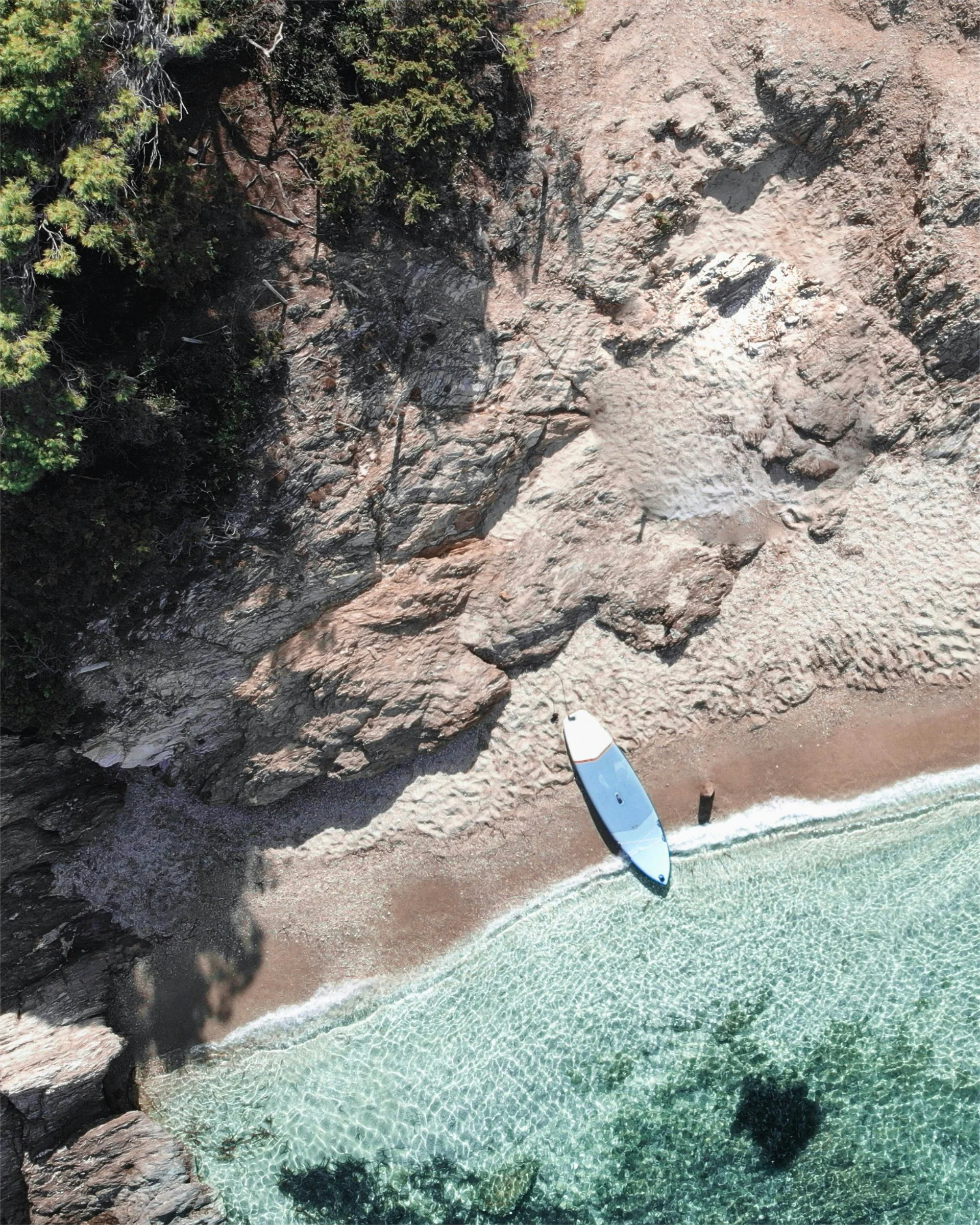 Aerial view of a paddleboard on a narrow strip of sandy beach surrounded by rocky coastline and clear turquoise waters.