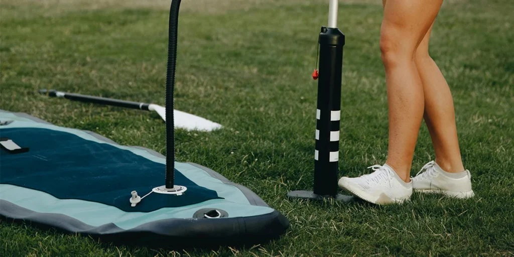 Close-up of inflatable paddleboard with manual pump on grass, alongside paddle and person's legs wearing sneakers.