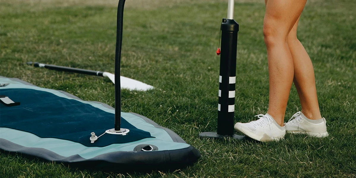 Close-up of inflatable paddleboard with manual pump on grass, alongside paddle and person's legs wearing sneakers.