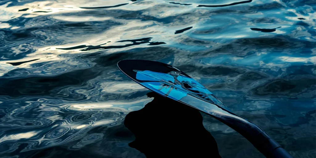 Close-up of a paddle with a reflective surface, resting on sparkling water with ripples and light reflections.