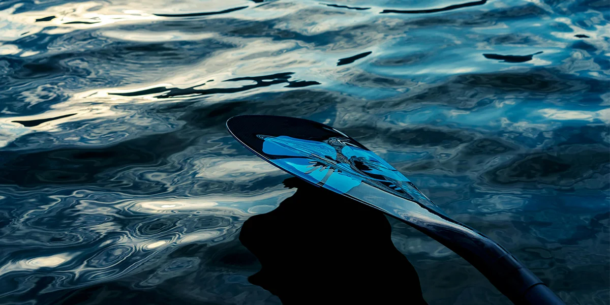 Close-up of a paddle with a reflective surface, resting on sparkling water with ripples and light reflections.