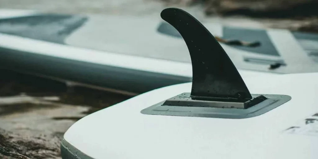 Close-up view of a black paddleboard fin attached to a white paddleboard, resting on wet ground with another paddleboard in the background.