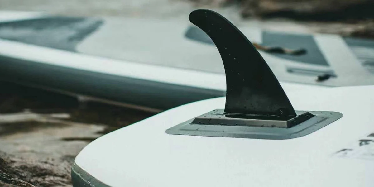 Close-up view of a black paddleboard fin attached to a white paddleboard, resting on wet ground with another paddleboard in the background.