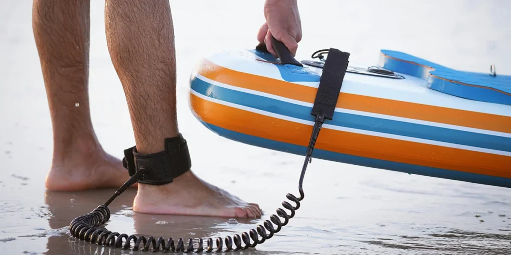 Close-up of a person standing barefoot on the beach, holding a paddleboard with a coiled ankle leash attached.