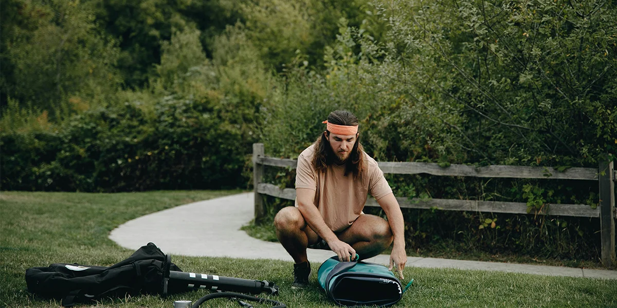 Man crouching on grass beside a park pathway packing equipment into a bag, surrounded by greenery and wooden fencing.