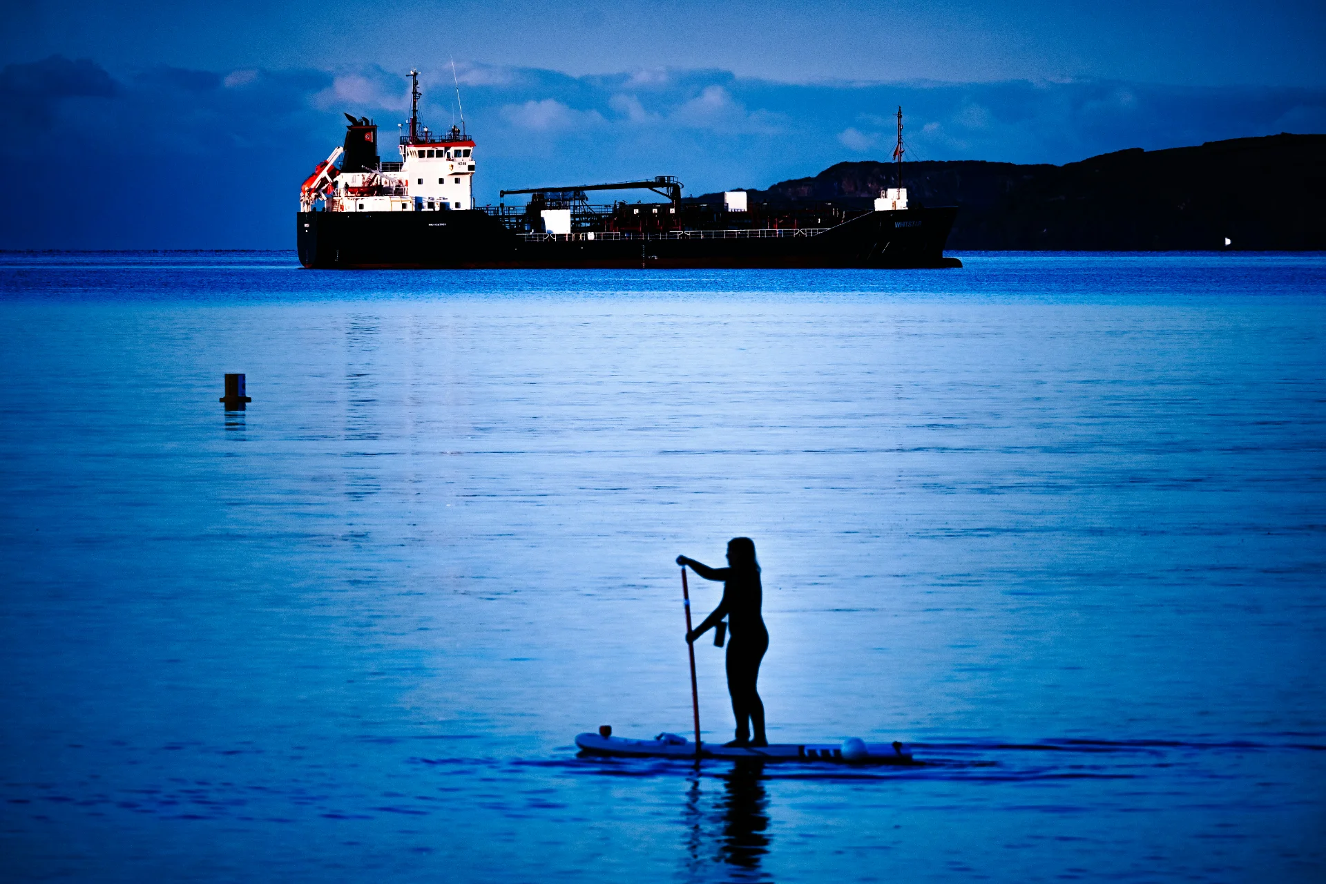 A silhouetted paddleboarder navigating calm waters near a large cargo ship and distant cliffs during dusk under a blue sky.