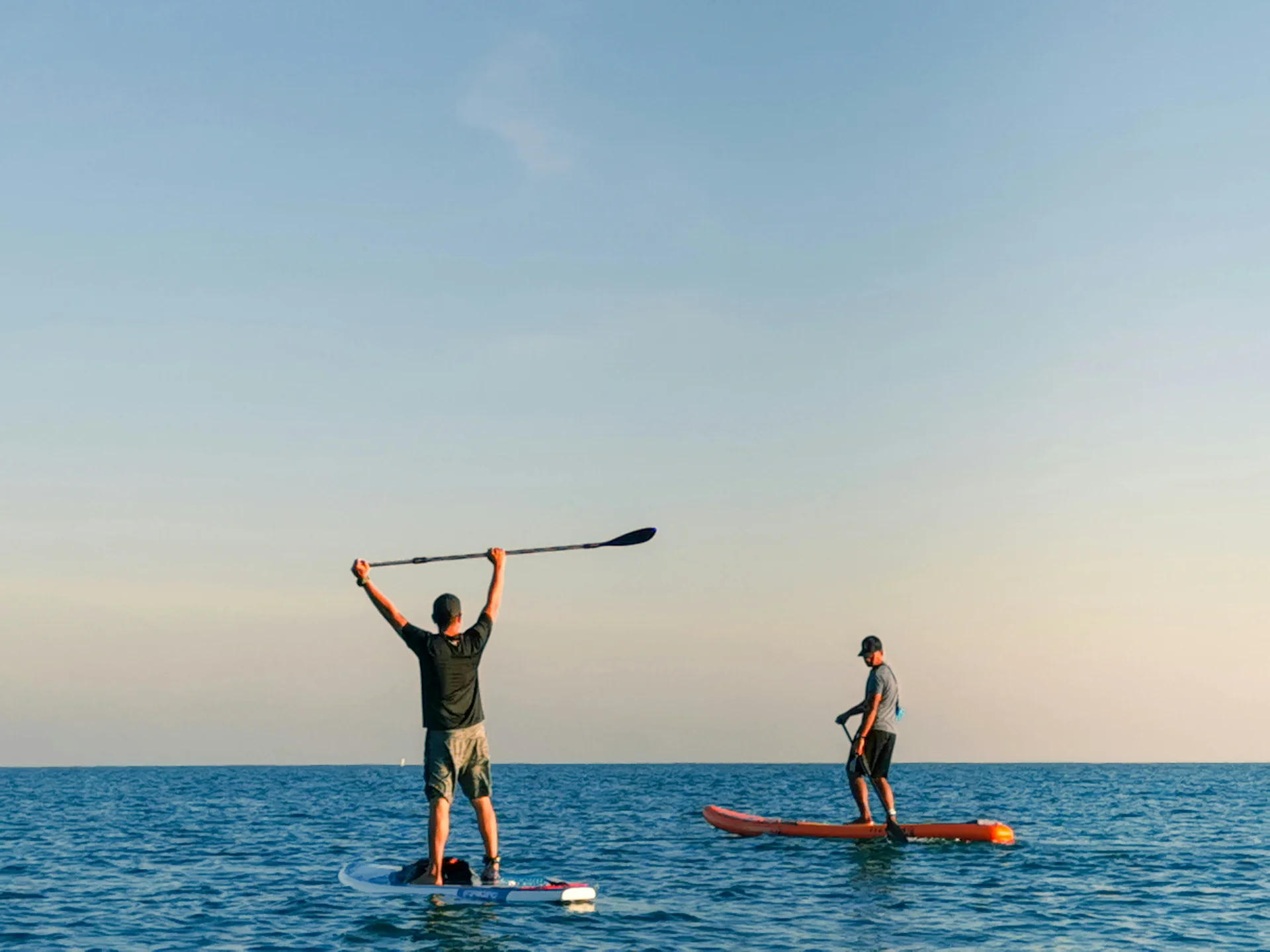 Two individuals paddleboarding on calm ocean waters during sunset, with one person raising their paddle triumphantly.