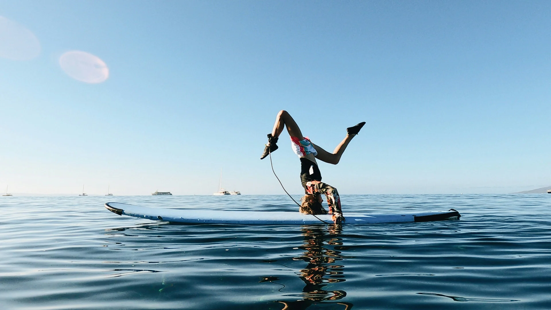 Woman performs an inverted yoga pose on a paddleboard surrounded by calm ocean waters, with sailboats and clear skies in the background.