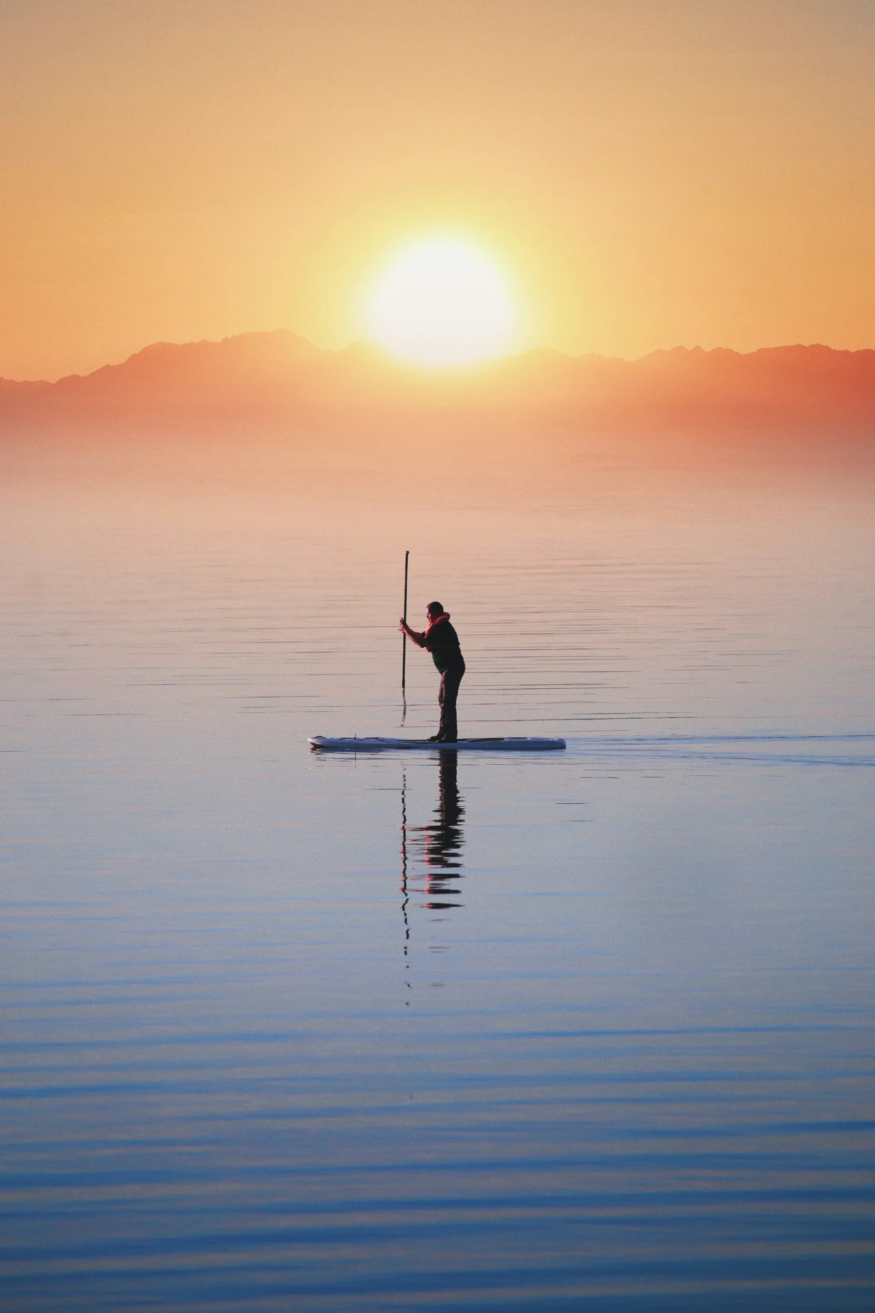 A person paddleboarding on still water during a serene sunrise, with warm orange tones, mountain silhouettes, and smooth reflections.