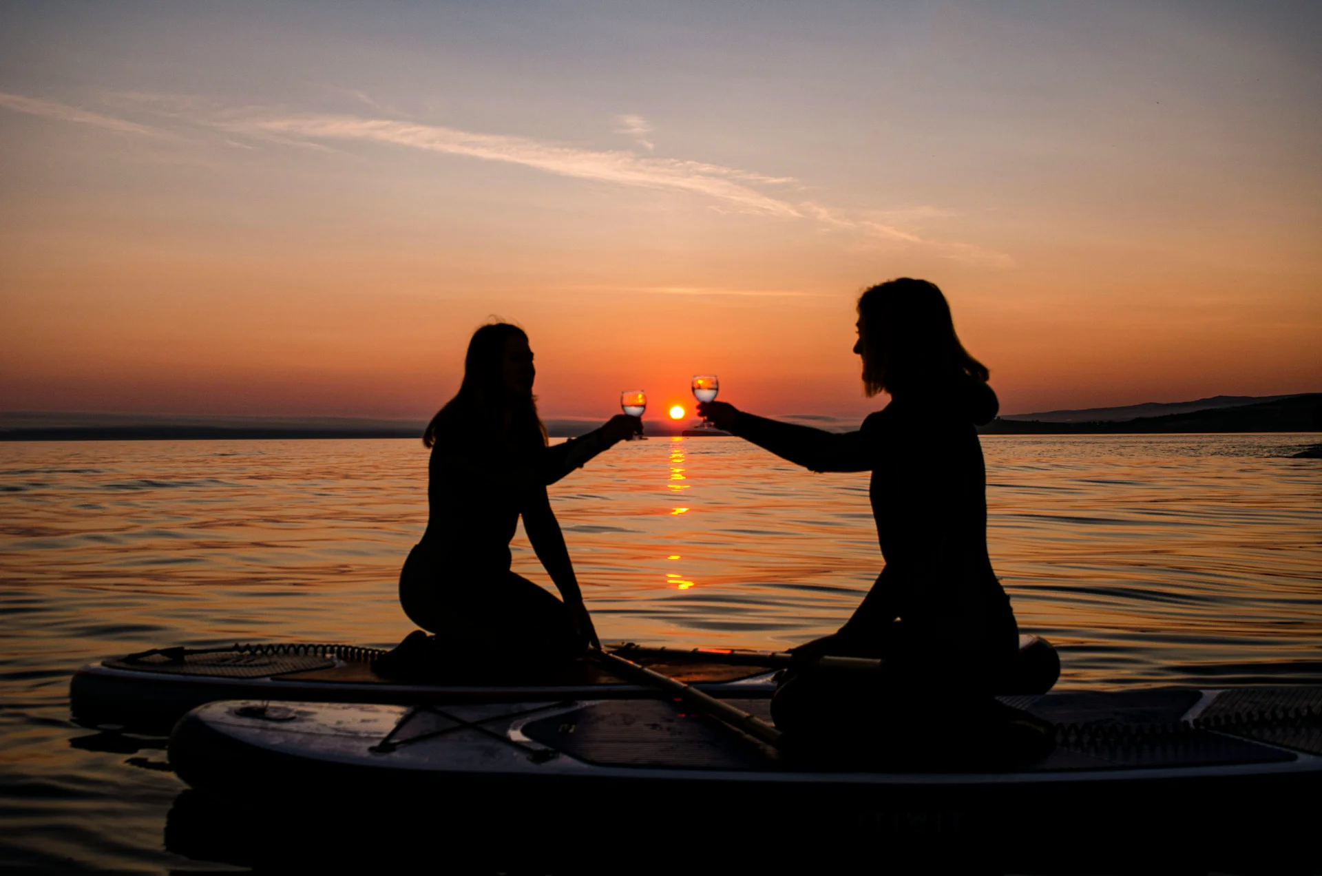 Two women on paddle boards toasting with drinks during sunset on calm water with golden and orange hues in the sky.