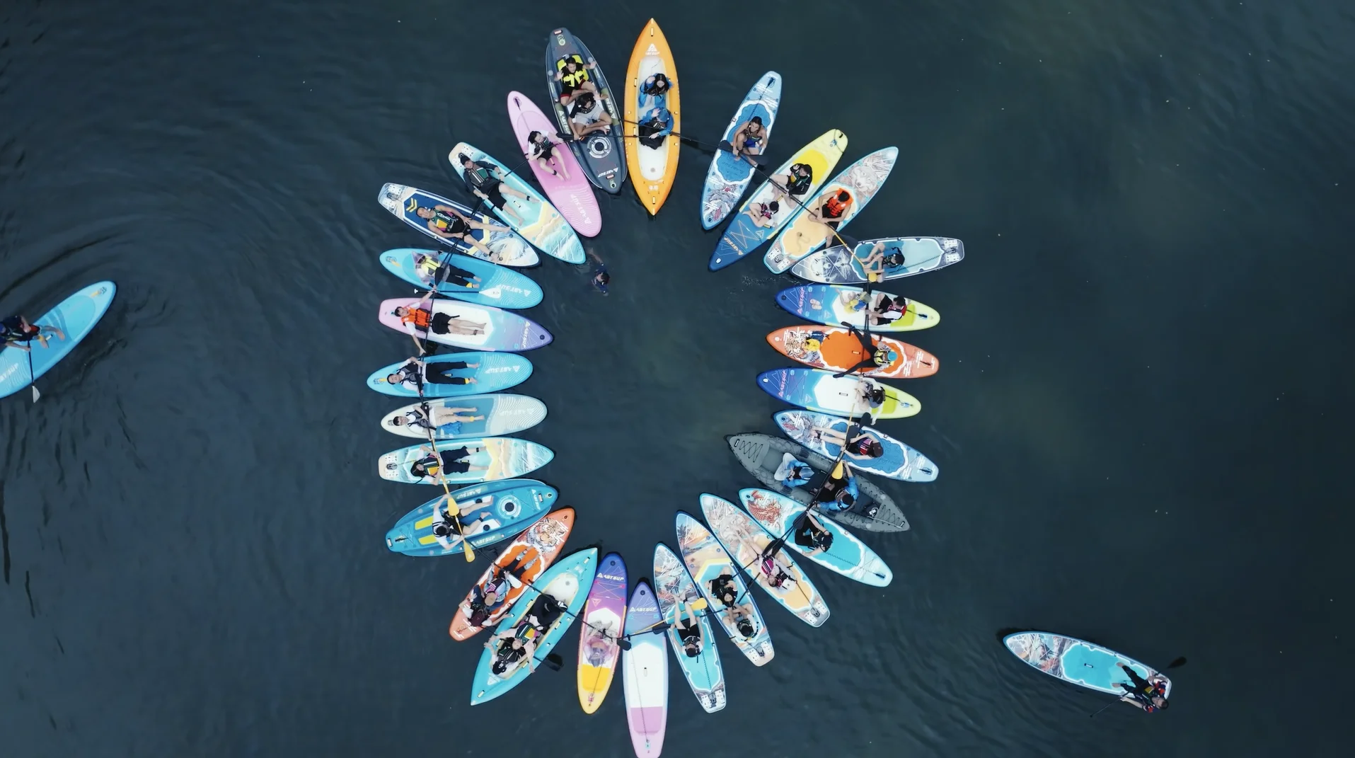 Aerial view of a group of paddleboarders creating a circular formation on calm water, with vibrant and colorful paddleboards.