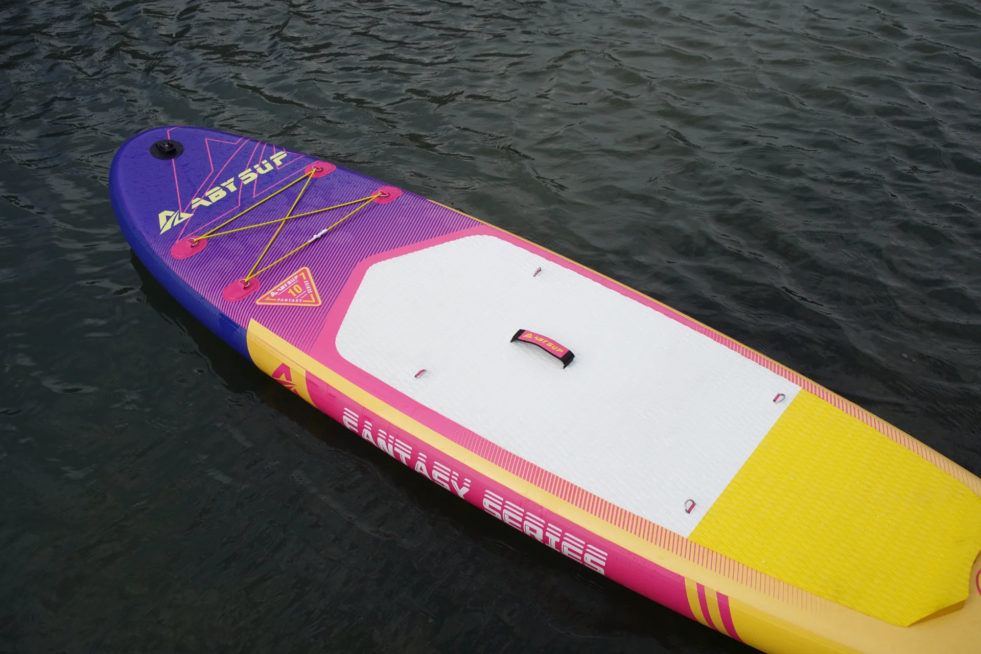 A vibrant purple, pink, yellow, and white paddleboard with a textured surface floating on calm water, featuring a central carry handle.