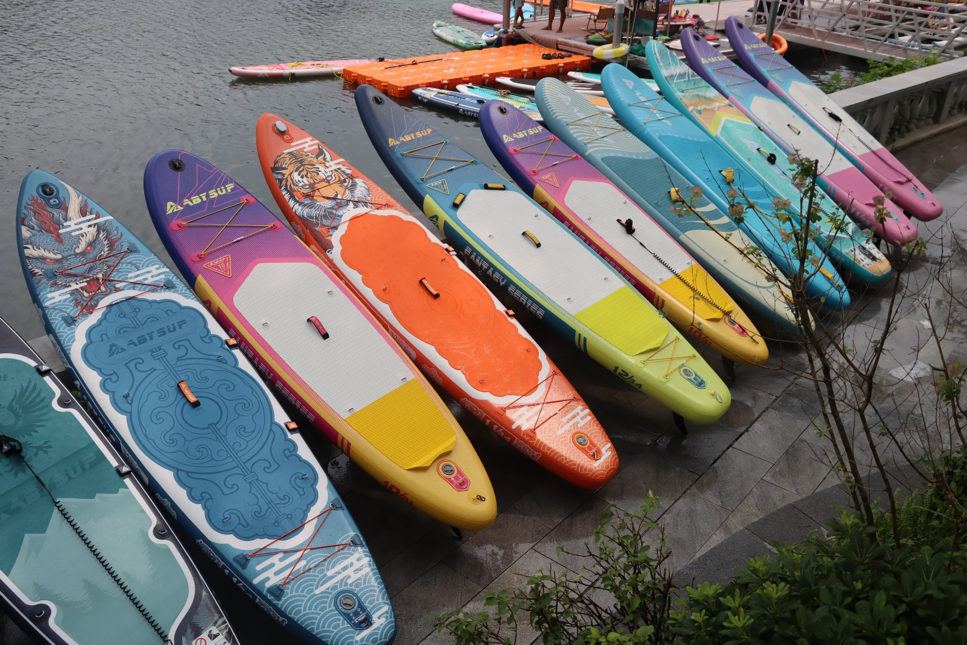 A row of colorful paddleboards with various designs, including tiger and dragon motifs, neatly arranged near a dock by the water.