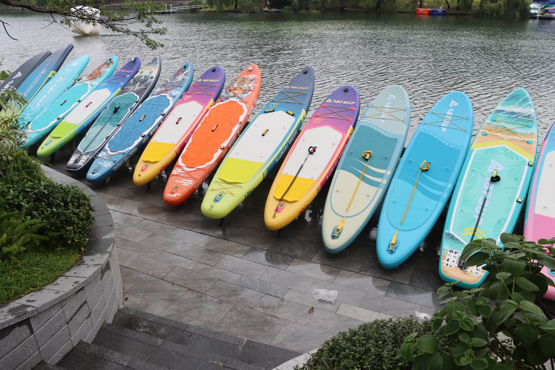 Brightly colored paddleboards arranged in a row on a stone-paved shoreline beside a calm lake with surrounding greenery.