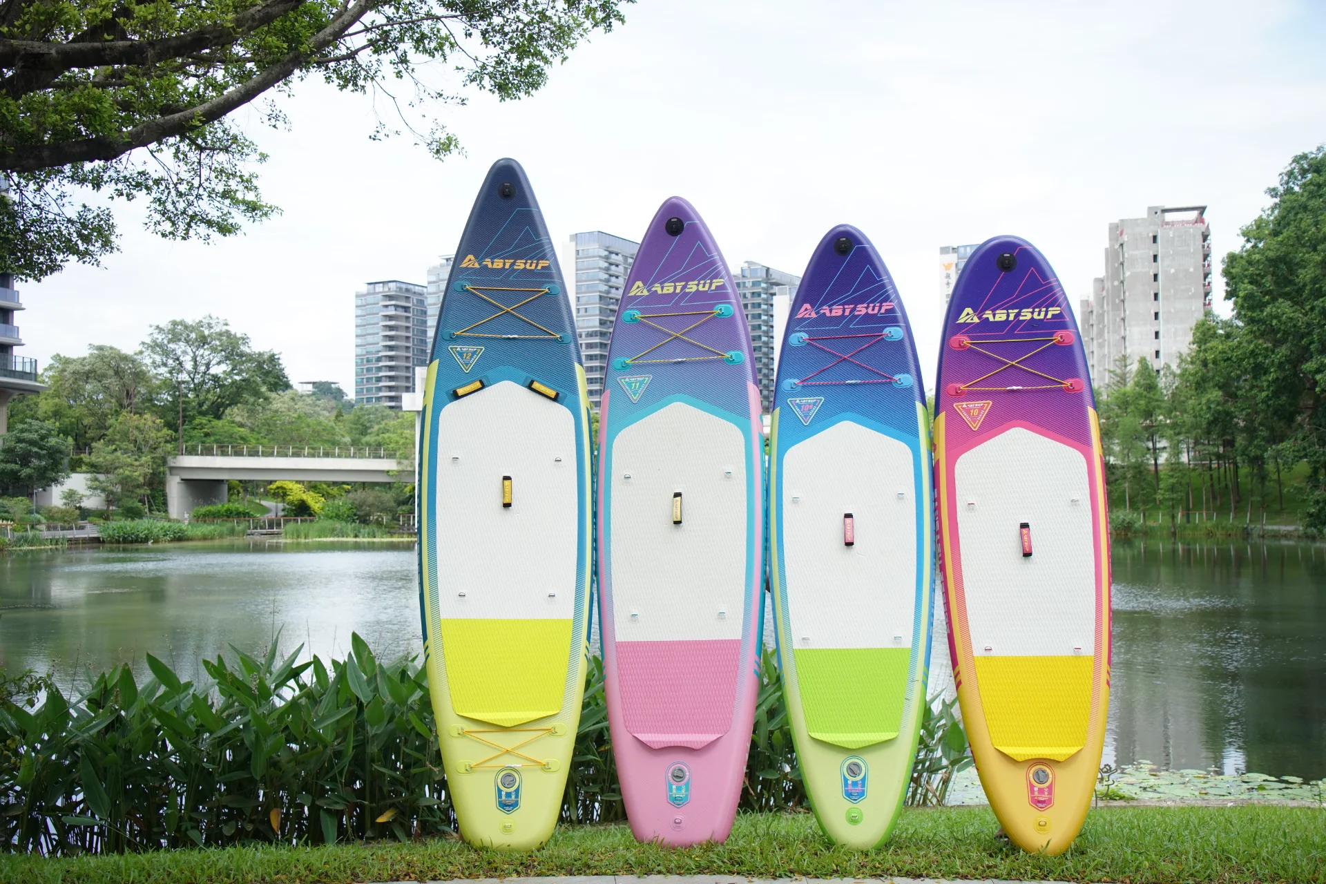 Four colorful paddleboards with vibrant designs standing upright near a serene lake surrounded by greenery and modern buildings.
