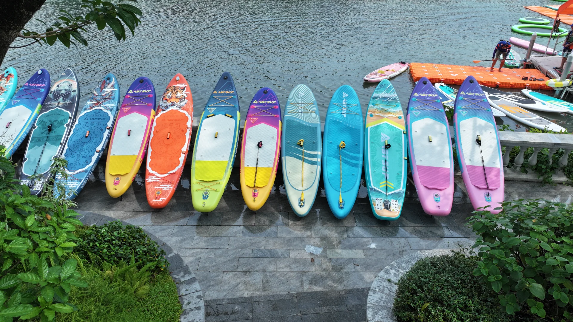 A row of brightly colored paddleboards with various designs lined up by the river, surrounded by greenery and a dock in the background.