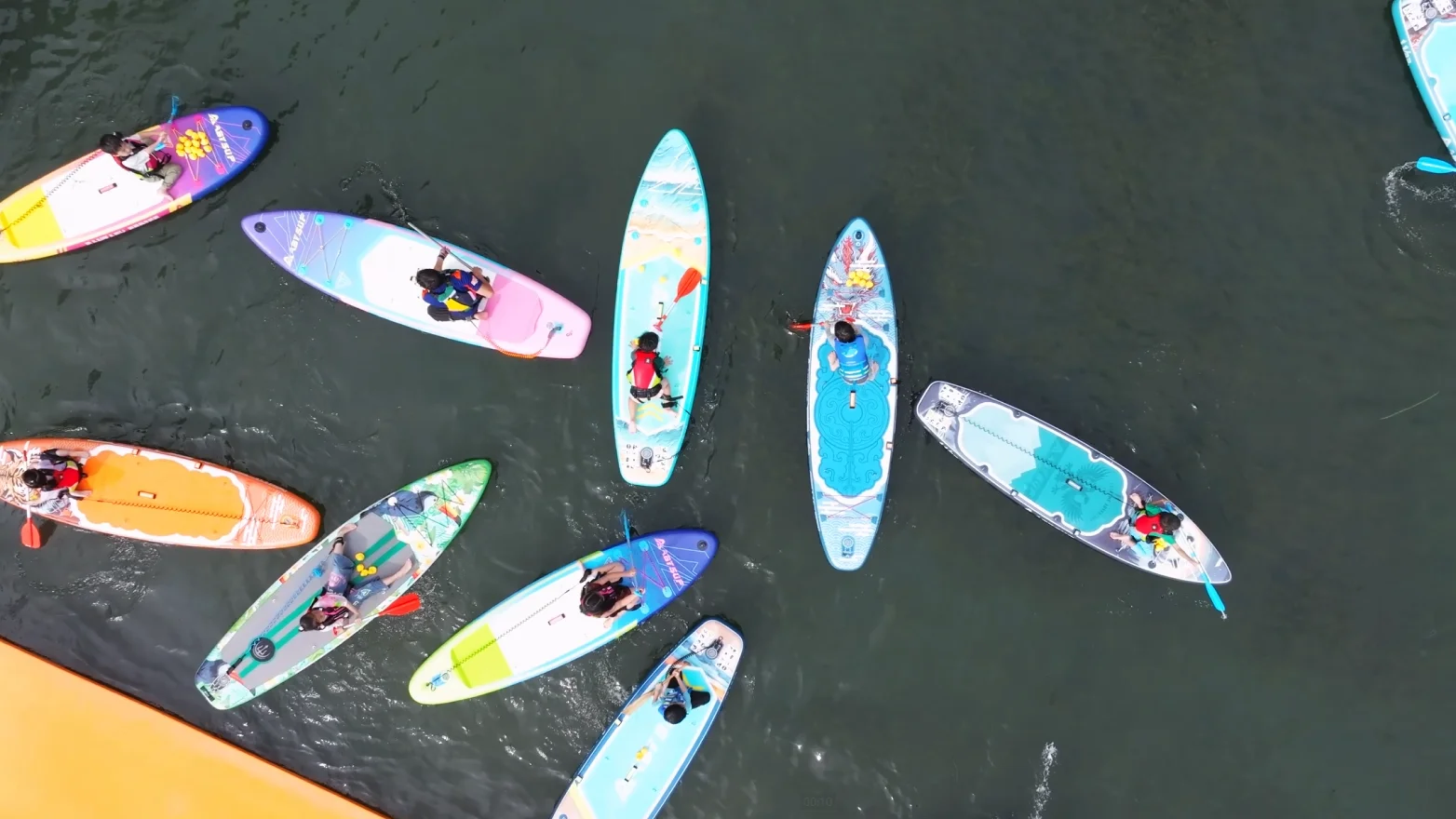 Bovenaanzicht van een groep paddleboarders die op kalm water drijven, kleurrijke boards en mensen die reddingsvesten dragen.