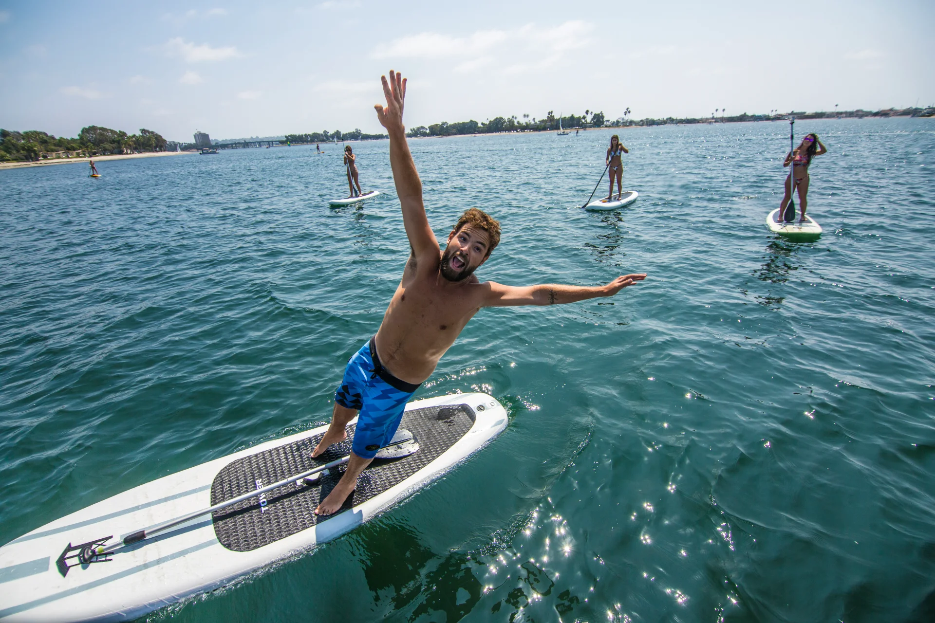 A man falling off a paddleboard in the foreground with three other individuals paddleboarding in the water under sunny skies.