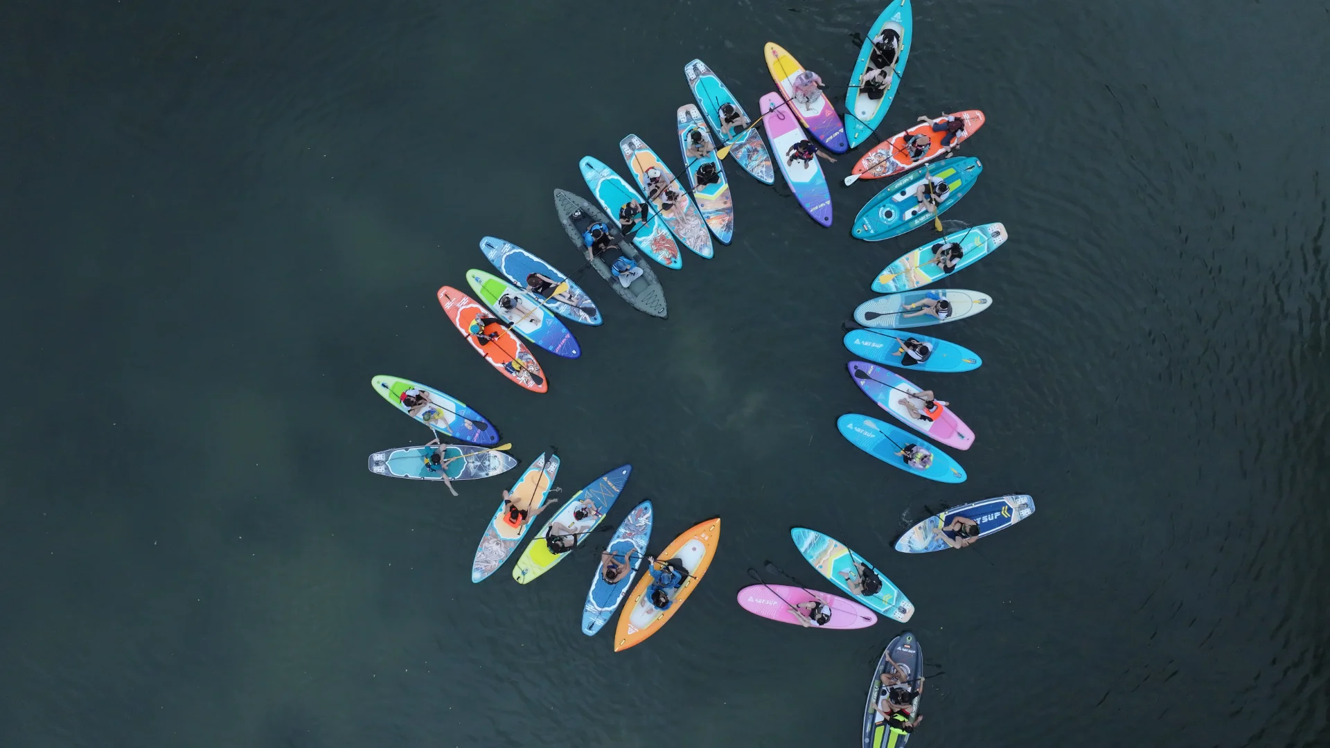 Aerial view of people on colorful paddleboards arranged in a circular formation on a calm lake.