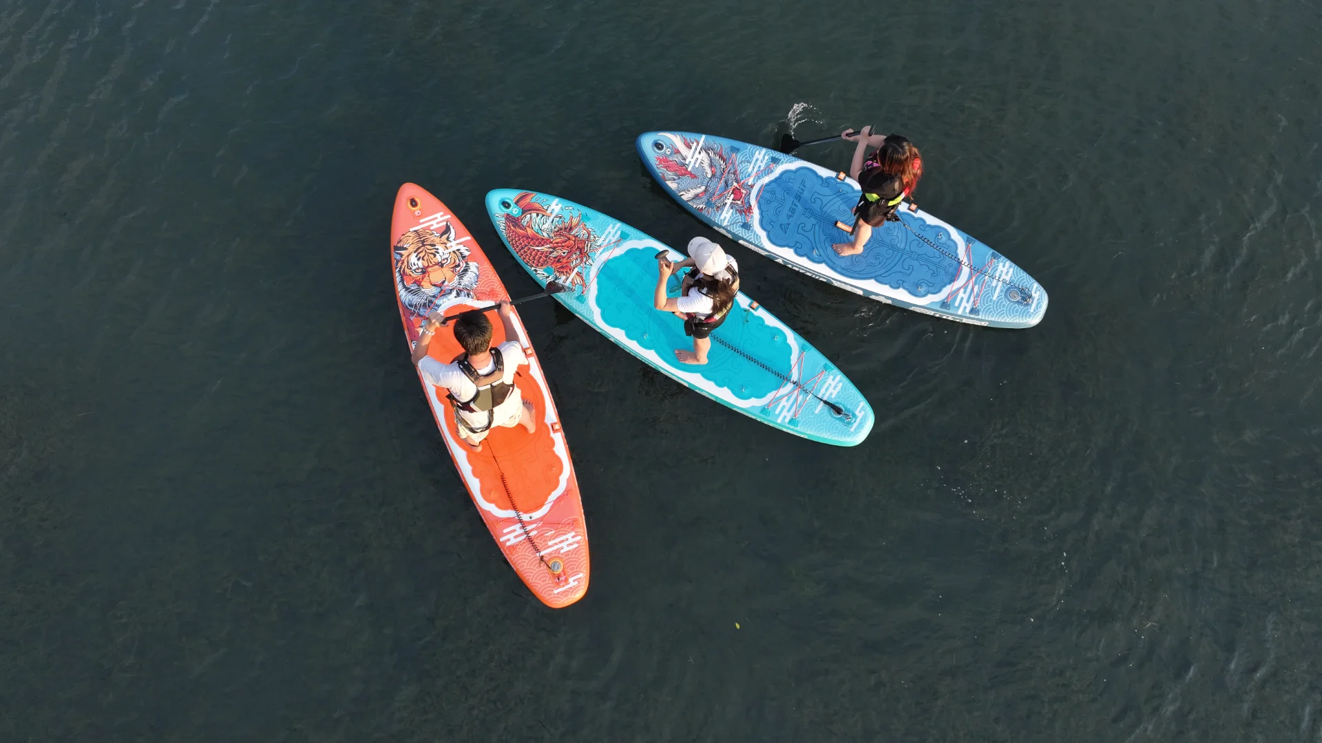 Aerial view of three paddleboarders on ornate boards floating on calm water, wearing life vests and holding paddles.