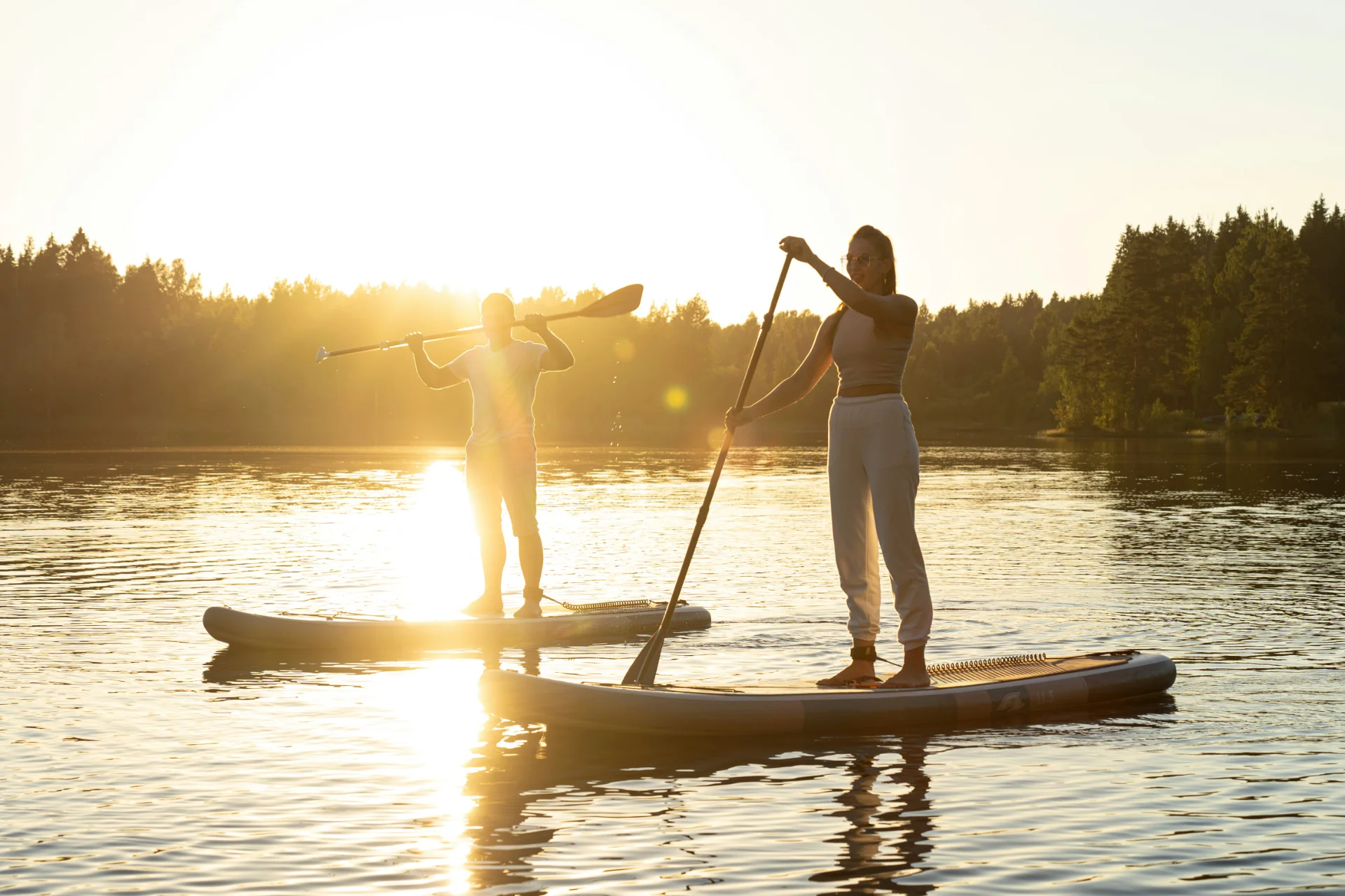 Two individuals on paddleboards during sunset, surrounded by calm waters and forested shoreline, enjoying outdoor activities.