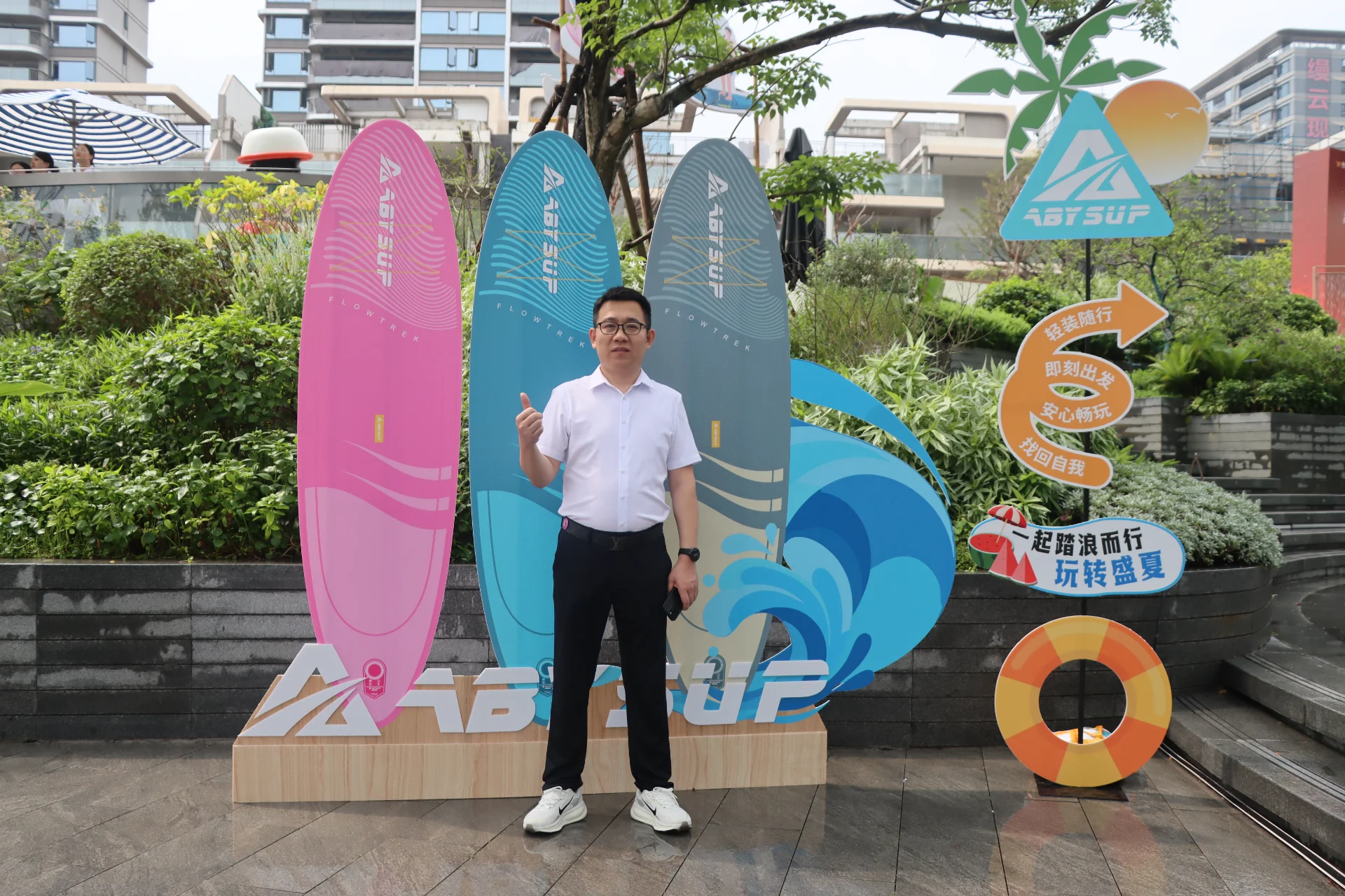 Man in white shirt and dark pants standing near vibrant surfboards at an outdoor event surrounded by greenery and promotional signs.