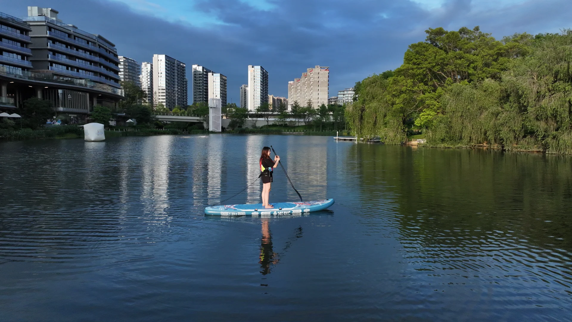 A woman paddleboarding on a calm lake surrounded by modern buildings and green trees under a cloudy blue sky.