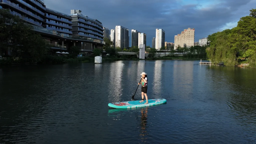 A woman wearing a white cap and life vest paddleboarding on a calm urban lake near modern buildings and lush trees at sunset.