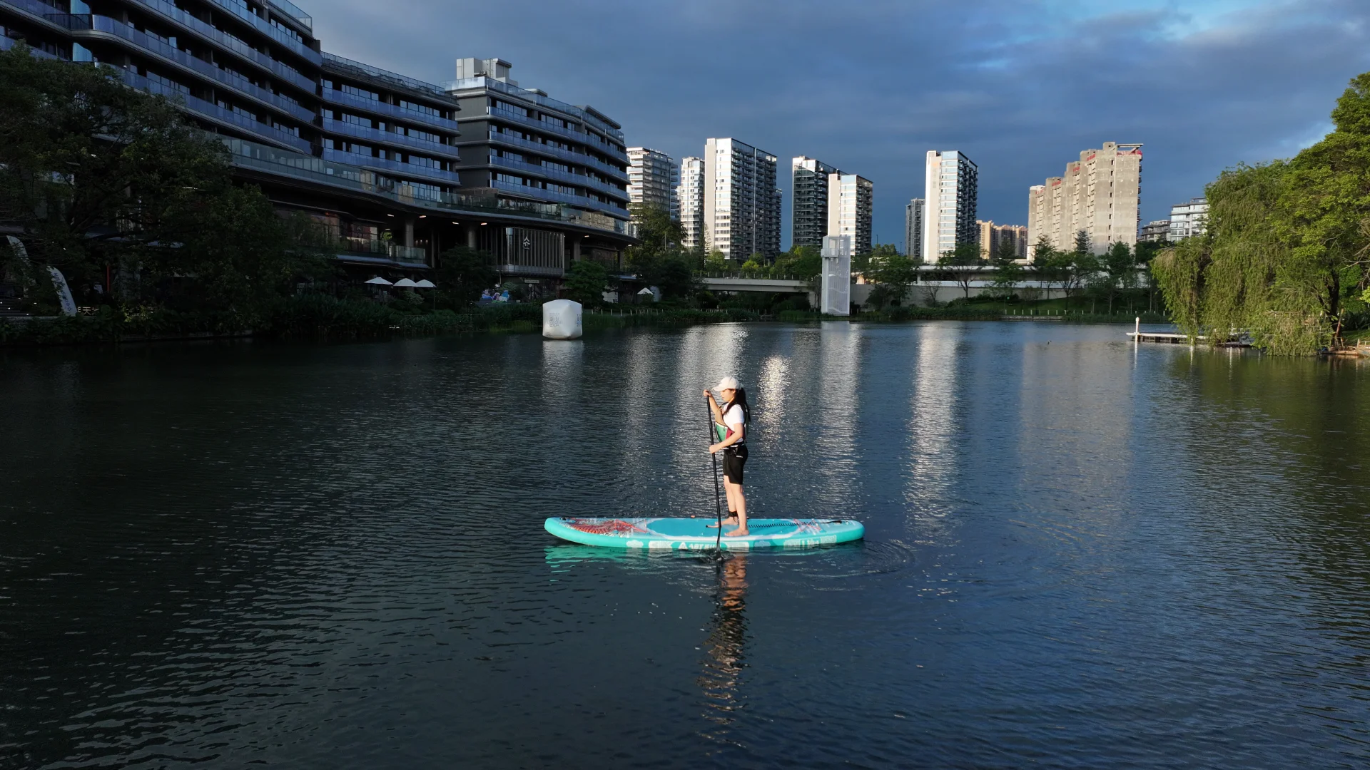 Woman standing on a paddleboard and paddling through a calm urban river surrounded by modern buildings, trees, and a bridge.