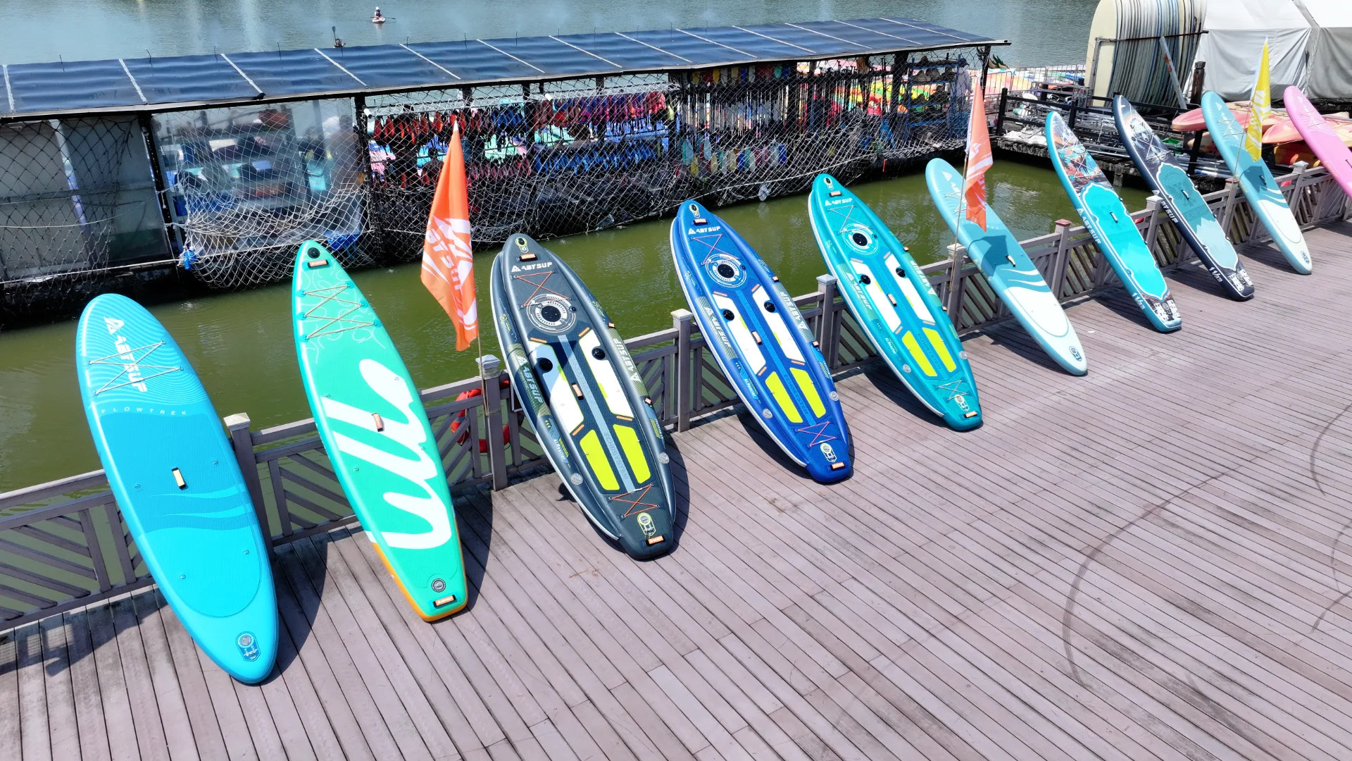 A row of colorful paddleboards displayed on a wooden dock near the water. An orange flag is in the middle.