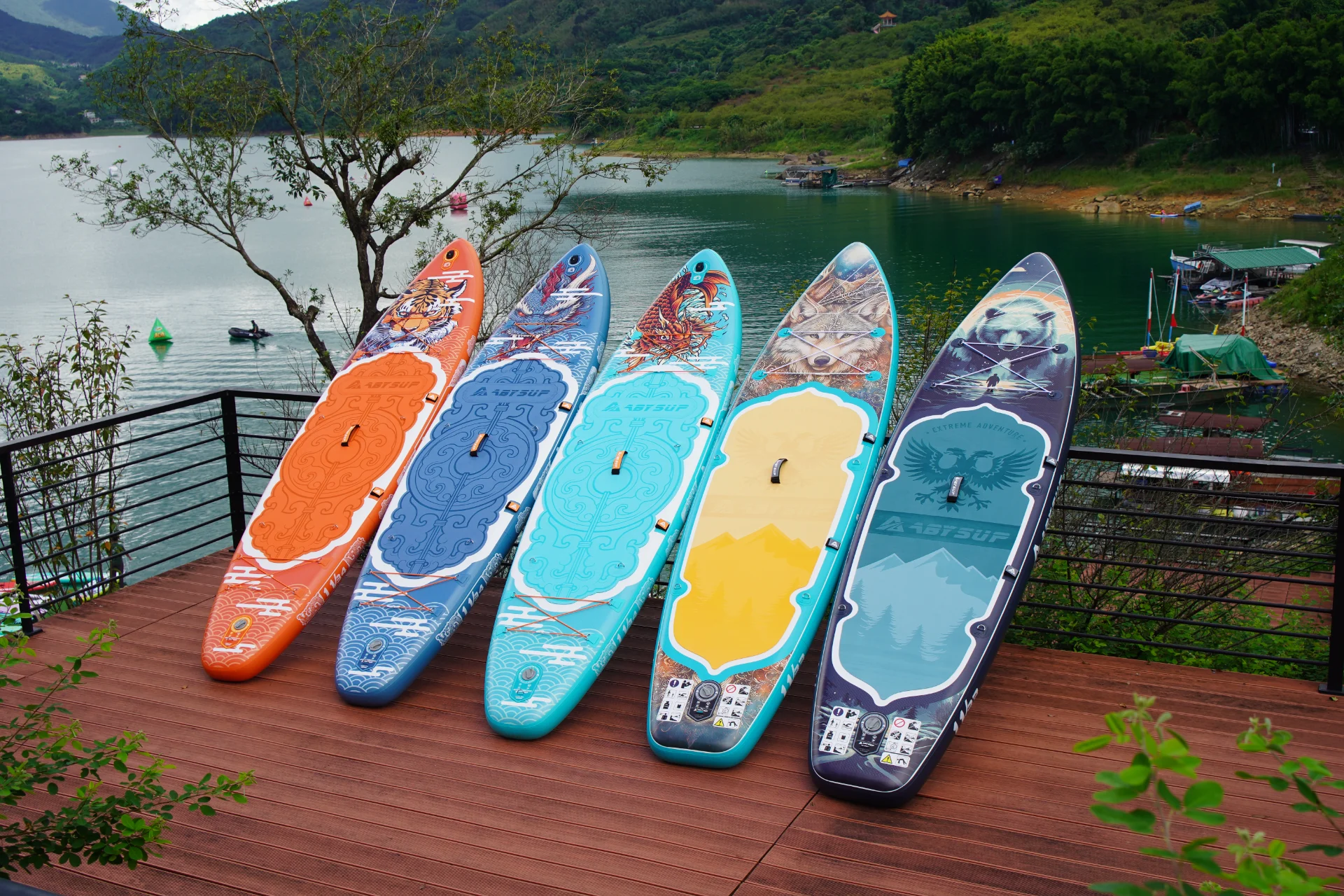 Five colorful and uniquely designed paddleboards arranged side-by-side on a wooden deck by a scenic lake surrounded by hills.