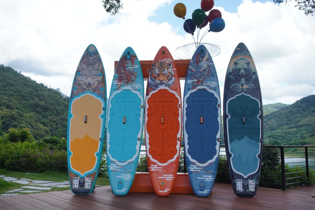 A row of five colorful paddleboards with artistic designs displayed outdoors on a wooden deck with a scenic mountain and lake background.