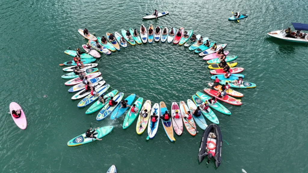 Aerial view of a group of paddleboarders forming a large circle in calm green-blue water with colorful boards and a nearby small boat.