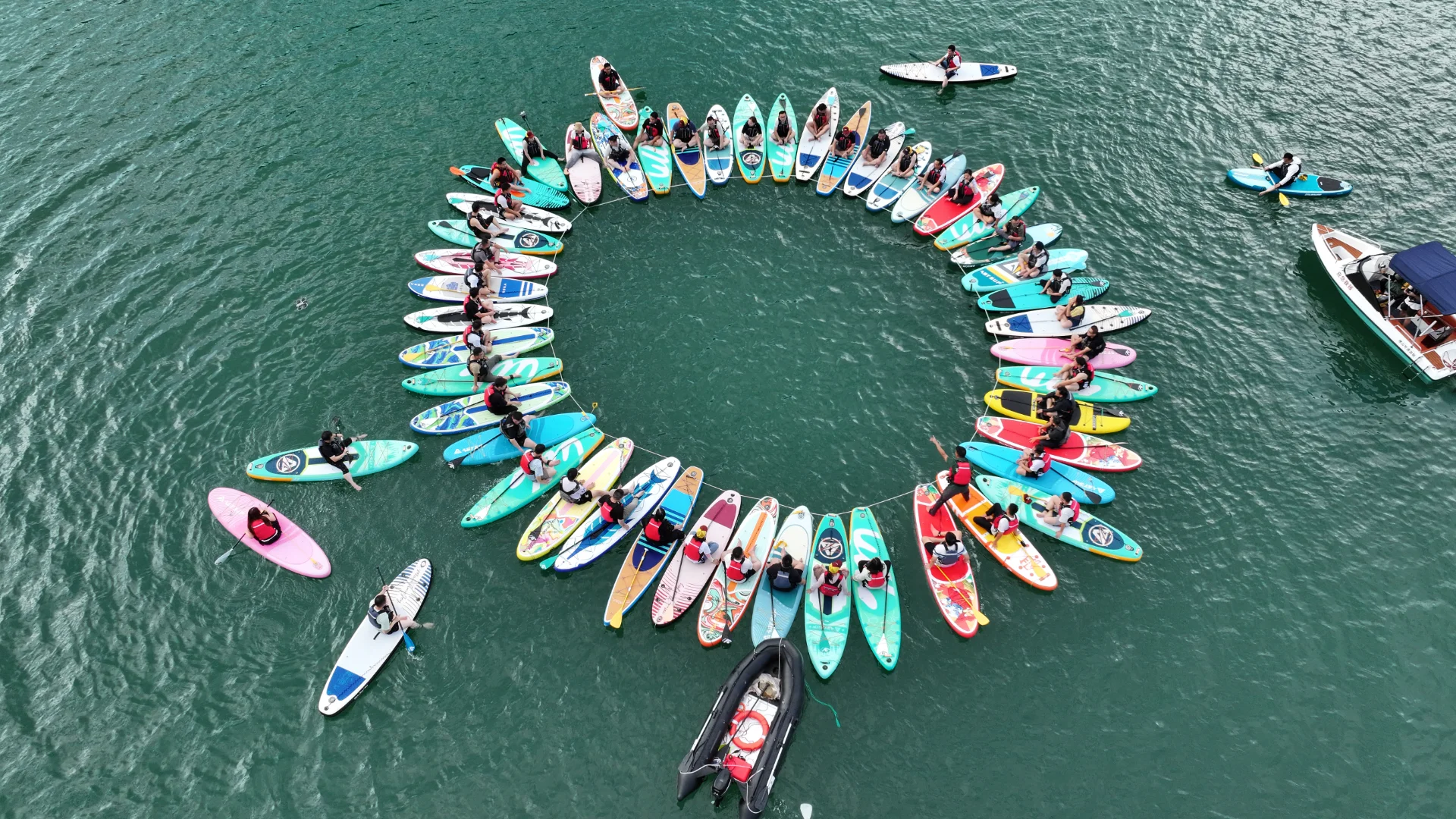Vista aérea de un grupo de personas sentadas en coloridas tablas de remo dispuestas en círculo sobre una tranquila masa de agua azul verdosa, con una pequeña lancha motora cerca.