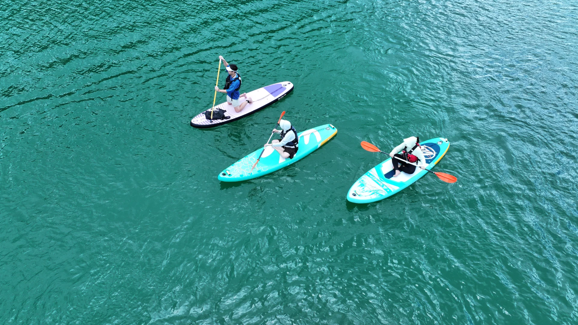 Three people paddling on a lake — one on a paddleboard and two in kayaks, with turquoise blue water surrounding them.