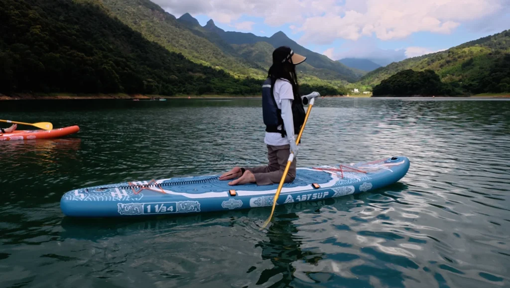 A person kneeling on a blue paddleboard with a paddle in hand, surrounded by a tranquil lake and green mountains in the background.
