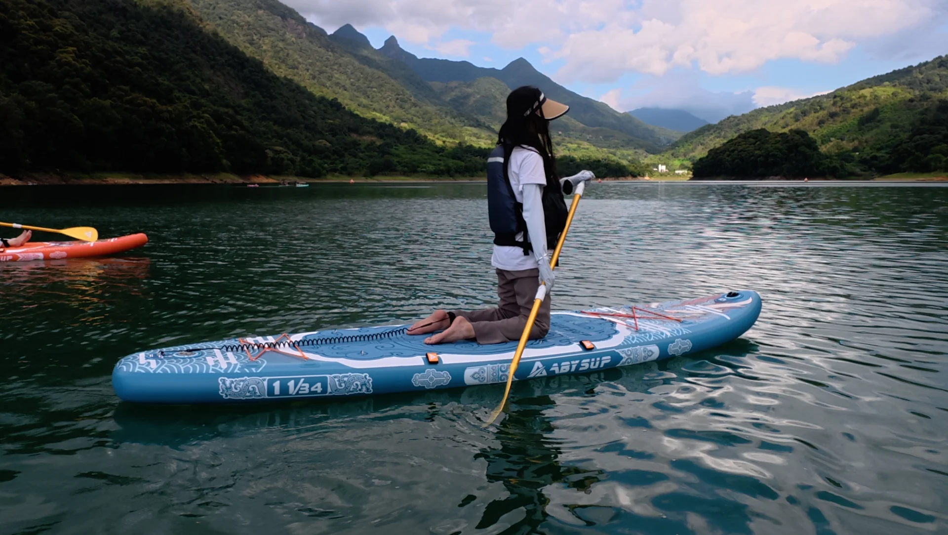 Een persoon knielt op een blauw paddleboard met een peddel in de hand, omringd door een rustig meer en groene bergen op de achtergrond.