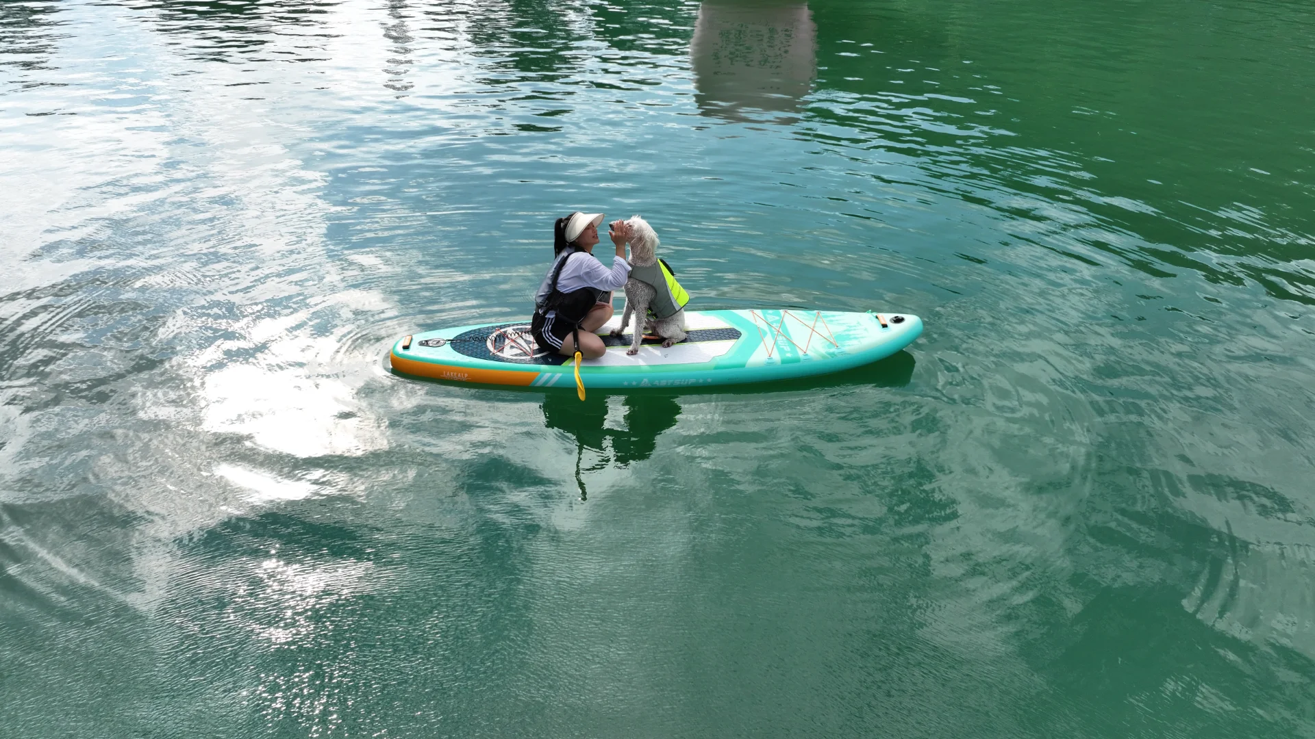 A woman sitting on a turquoise paddleboard on a calm green lake, interacting with her dog wearing a life jacket.